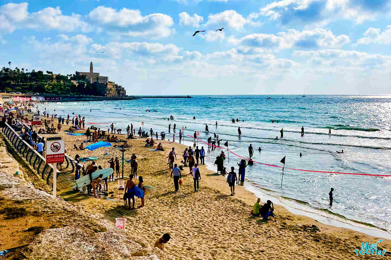 Crowded sandy beach with people swimming and relaxing by the sea, with an old town and lighthouse in the background