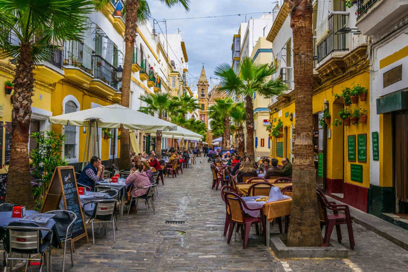 Lively Cadiz street showing a mix of traditional and modern architecture, with people enjoying outdoor dining under tall palm trees