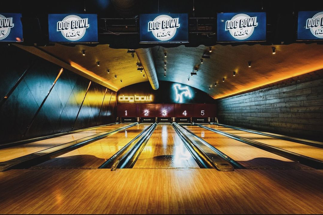 An atmospheric bowling alley with a distinctive, modern design featuring neon-lit lanes numbered one to five and the 'Dog Bowl' logo overhead