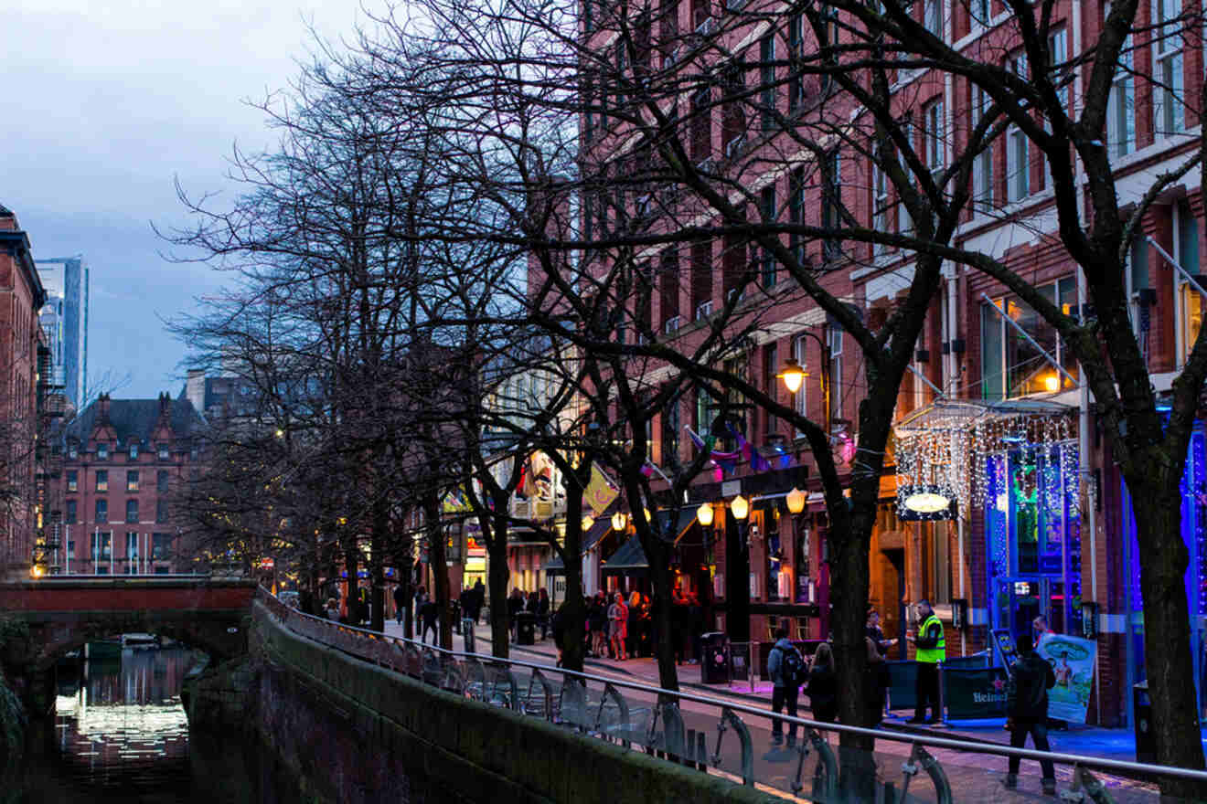 Evening view of Manchester's vibrant nightlife scene with illuminated street lights reflecting on a canal, surrounded by bustling walkways and buildings