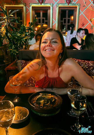 The writer of the post in a red top enjoys a meal with a glass of white wine at an elegantly decorated restaurant with a warm, ambient lighting