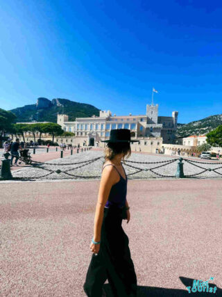 Author of the post in a casual outfit and hat walking across the Palace Square in Monaco, with the Prince's Palace in the background on a sunny day