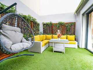 A vibrant outdoor lounge area at Family Aparthotel with yellow sofas, a wicker swing chair, and a coffee table, set on artificial grass and surrounded by green plant-covered walls