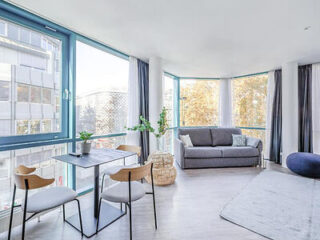 A bright and airy room at Limehome Köln Friesenplatz with a comfy gray sofa, dining area, large windows, and contemporary décor