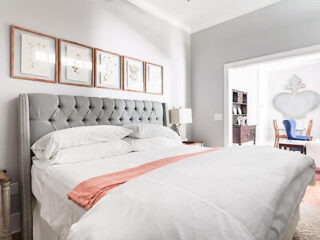 Elegant bedroom with a tufted gray headboard, framed botanical prints on the wall, and a soft coral throw over crisp white bedding