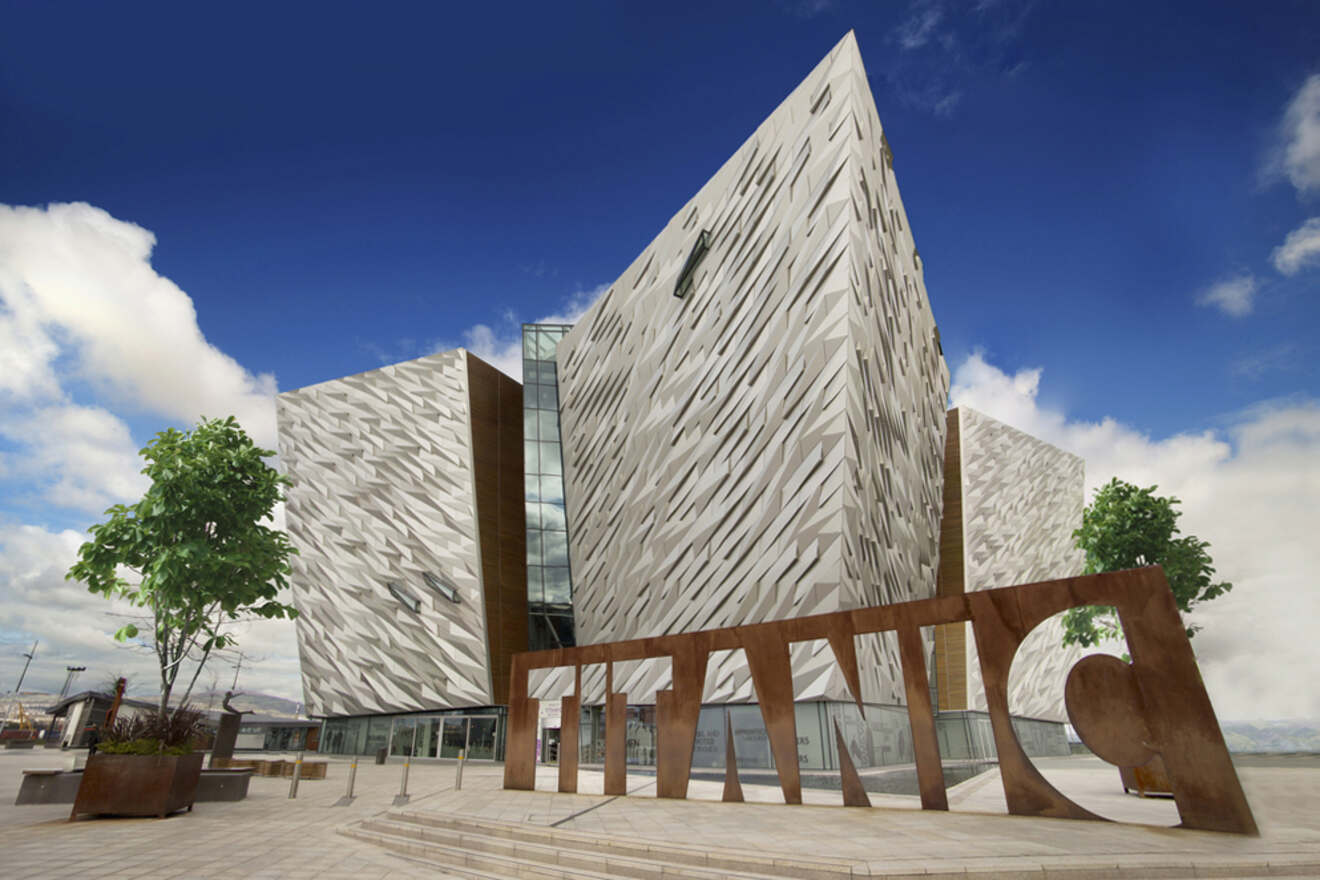 Modernistic façade of the Titanic Belfast museum, featuring angular, metallic shards that mimic the hull of the iconic ship, set against a backdrop of blue sky and clouds.