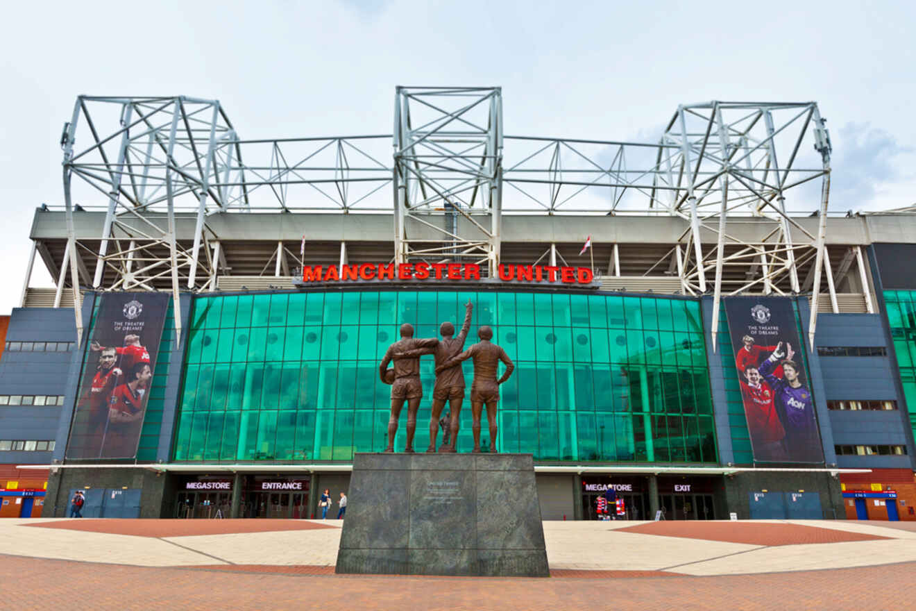 The front facade of Old Trafford, the Manchester United Football Stadium, with the statue of the 'United Trinity' in the foreground under a bright blue sky