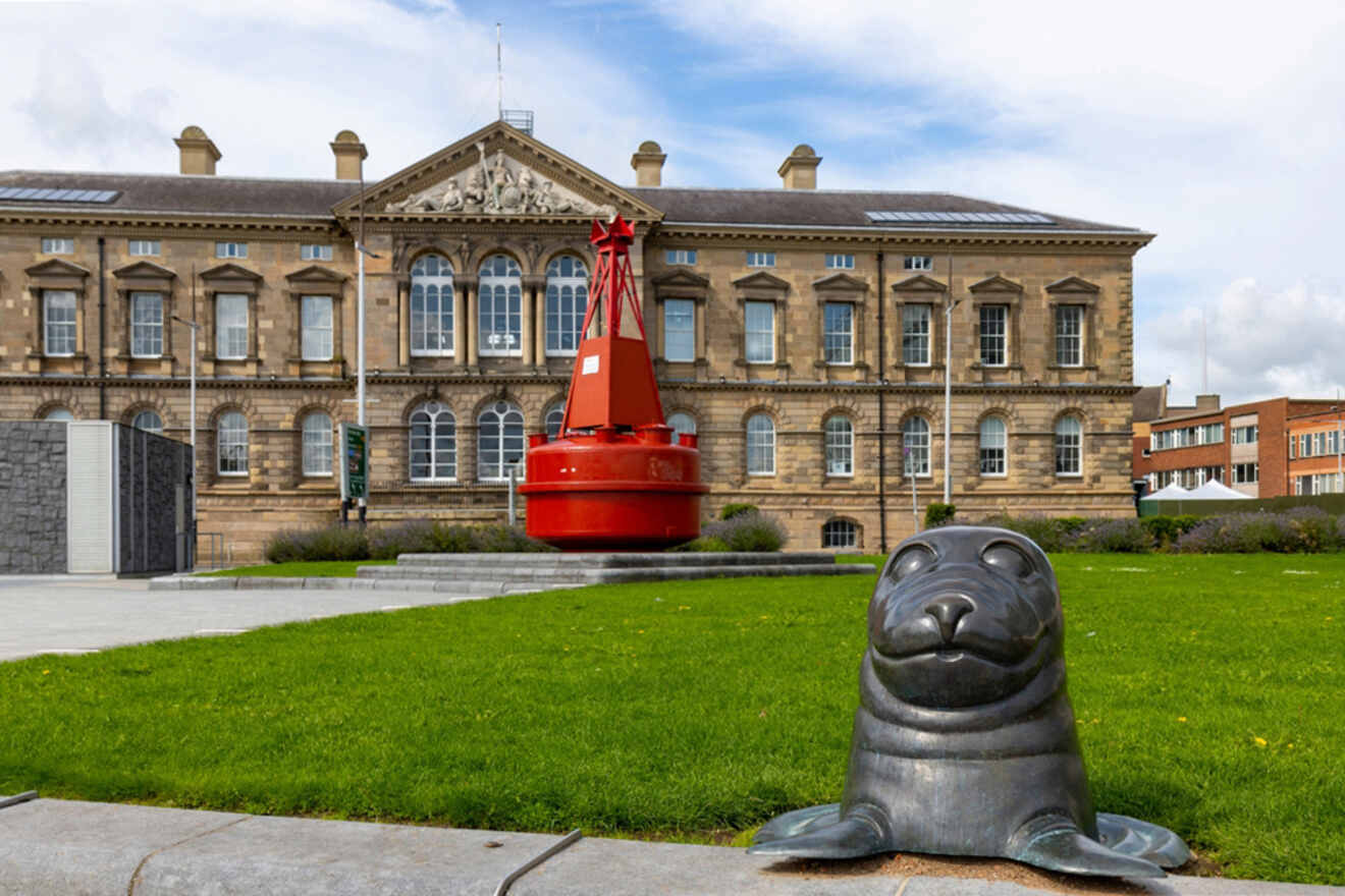 Exterior view of the Custom House in Belfast with a red navigational buoy and a seal sculpture in the foreground on a day with a mix of clouds and sunshine