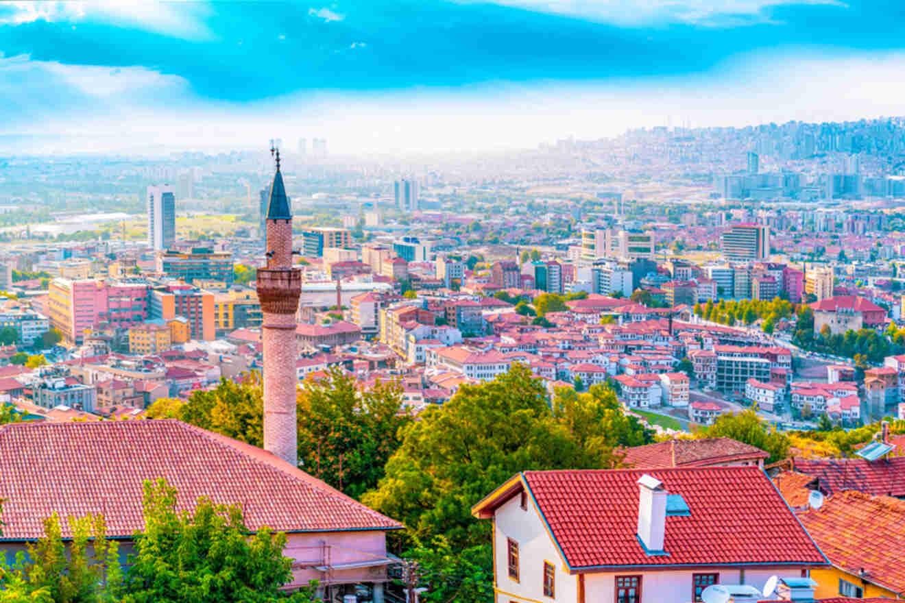 A captivating view over Ankara, Turkey, with the foreground showcasing a historic minaret and red-roofed buildings against a sweeping cityscape and hazy blue skies.