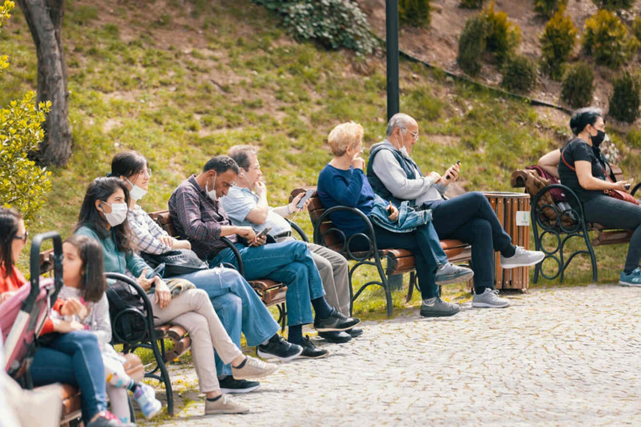 People sitting on benches in a park, some wearing masks, indicating a casual outdoor setting.