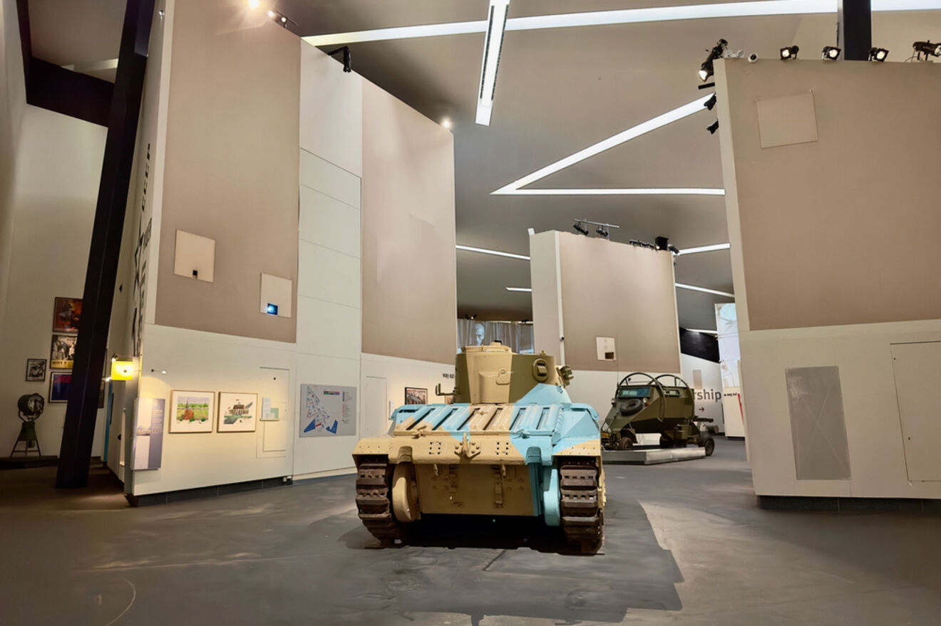 Interior of a spacious museum displaying military vehicles, including a prominent light blue tank, with white walls and modernistic lighting fixtures.
