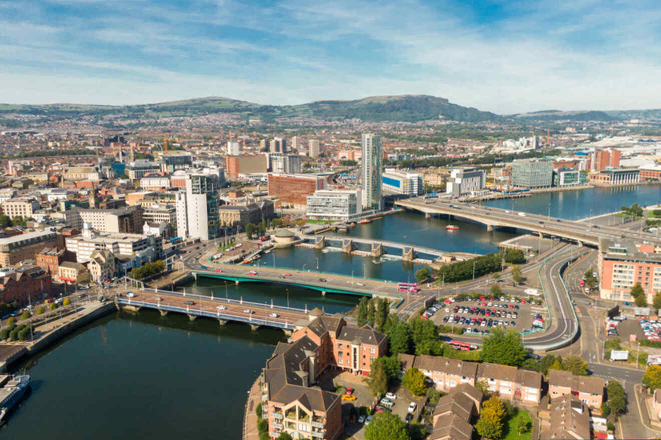 Aerial view of Belfast with the River Lagan flowing through the city. The image captures a clear day with a mix of modern buildings and historic architecture, traffic on the bridges, and a backdrop of green hills.