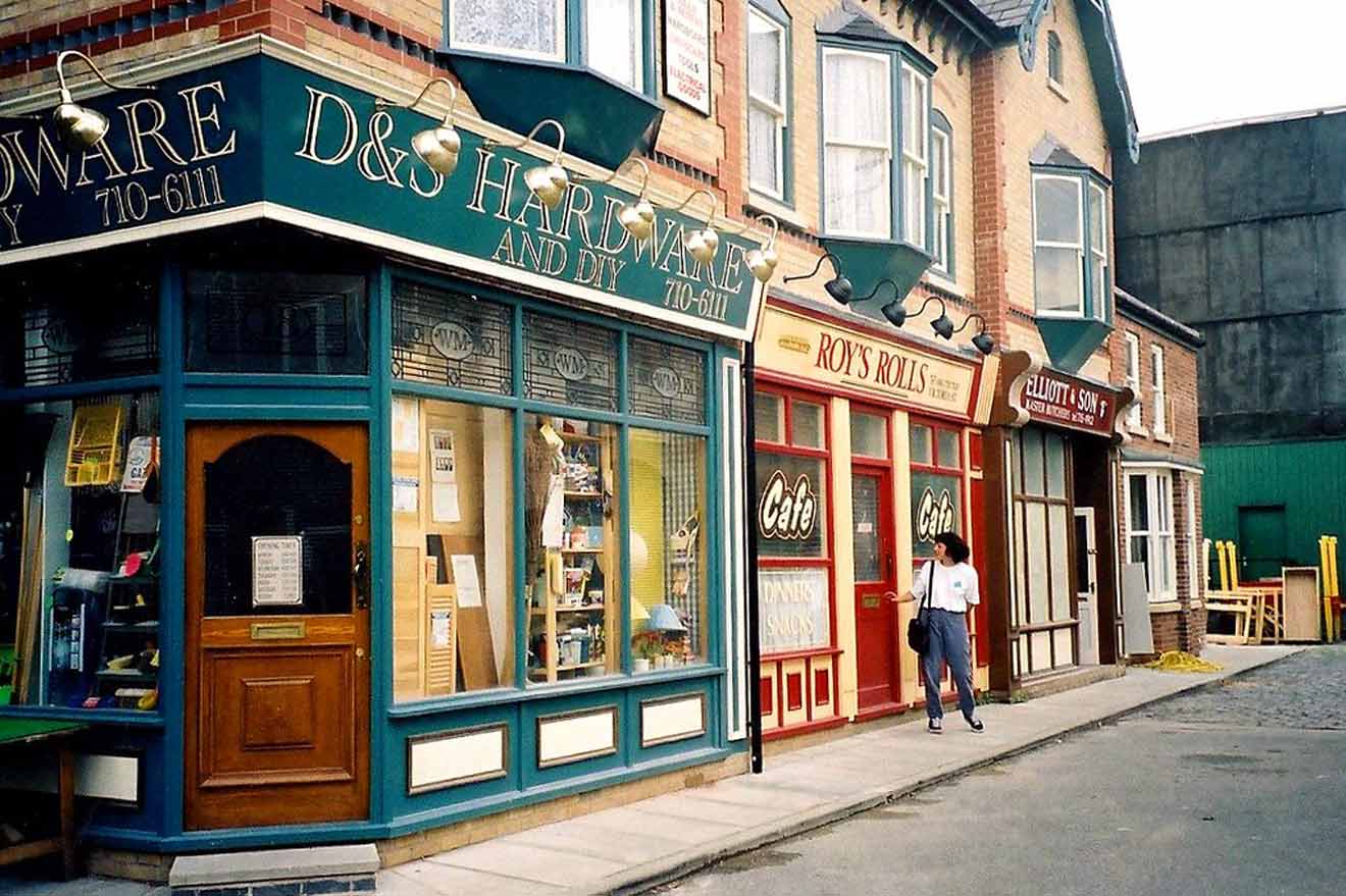 Quaint street scene from The Coronation Street Experience in Manchester, showing a row of traditional British shops including a hardware store and a café named 'Roy's Rolls