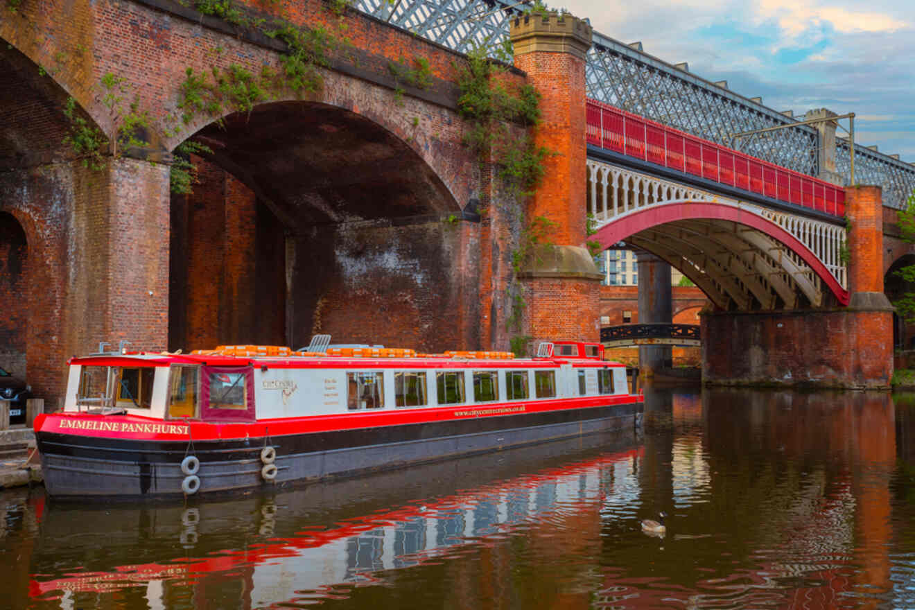A red and white canal boat named 'Emmeline Pankhurst' docked on the calm waters of a Manchester canal with arched brick bridges and urban buildings in the background