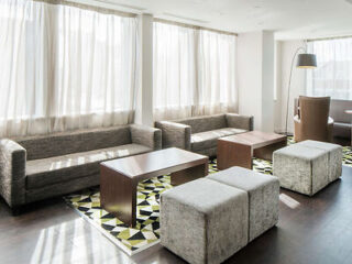 Minimalist hotel lobby with floor-to-ceiling windows, natural light, grey square sofas, wooden tables, and a geometric patterned rug