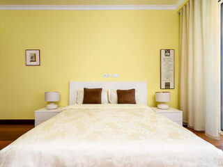 "Stylish bedroom with a bright yellow wall, a large bed with a cream-colored comforter, dark brown cushions, and minimalist decor