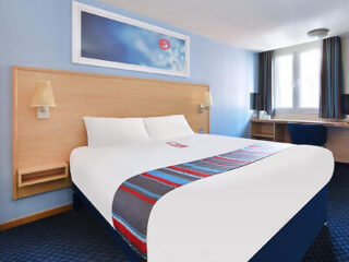 Hotel room with a white double bed featuring a blue and red runner, wooden headboard, framed artwork above the bed, and a small bedside lamp