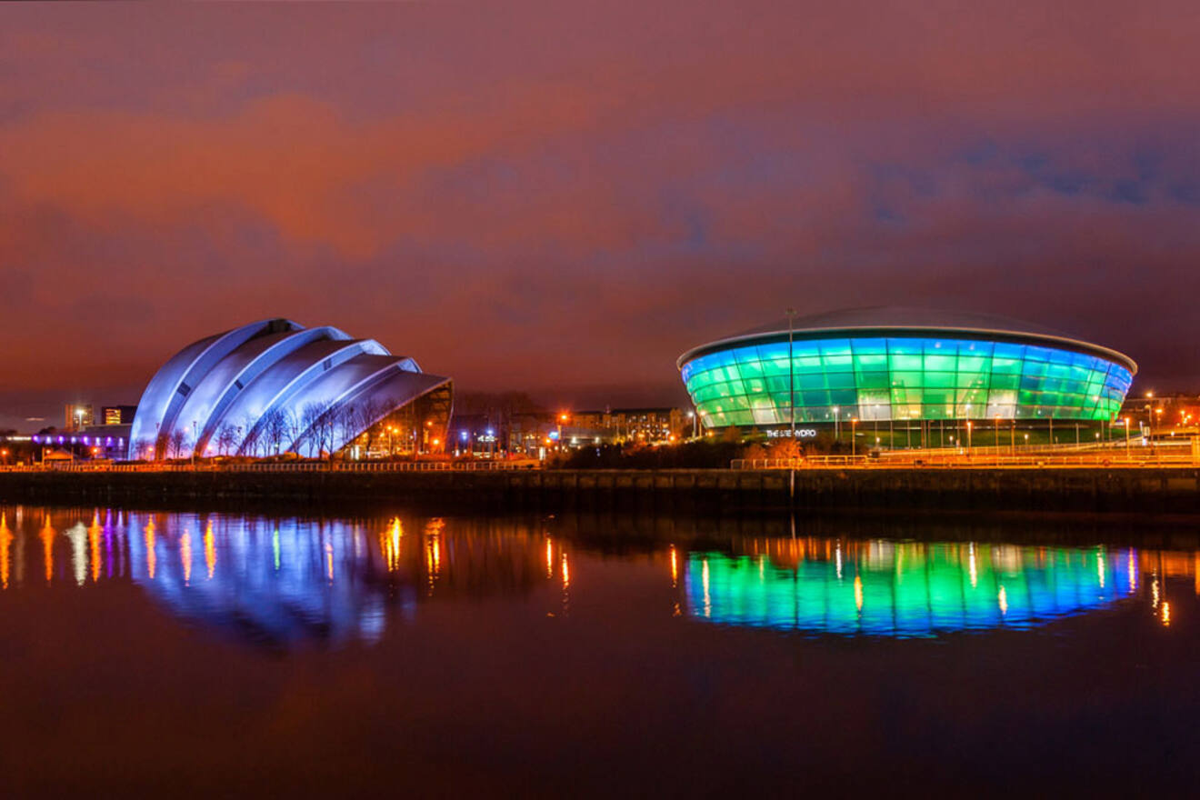 Night view of the lit-up, modern architecture of Finnieston in Glasgow, with the SSE Hydro's colorful illumination reflecting on the River Clyde, highlighting the area's urban vibe