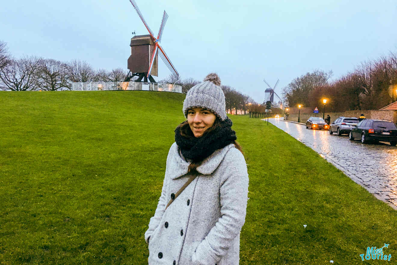 Author of the post in a winter hat and coat standing before a grassy slope with historic windmills in the background, on a wet cobblestone path in Bruges
