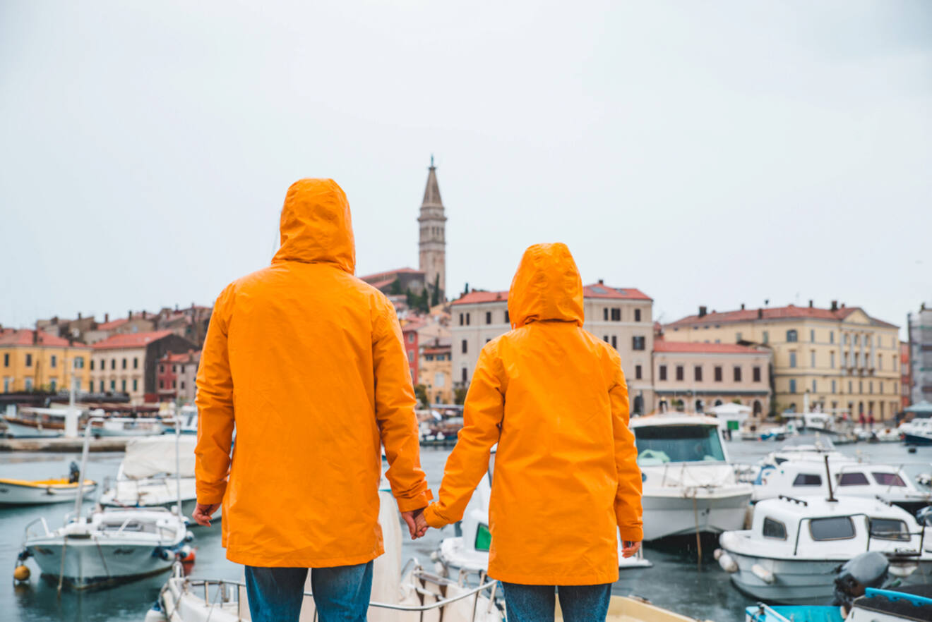 Two people in orange raincoats holding hands and looking towards a scenic view of Rovinj's waterfront and bell tower, with moored boats on a cloudy day