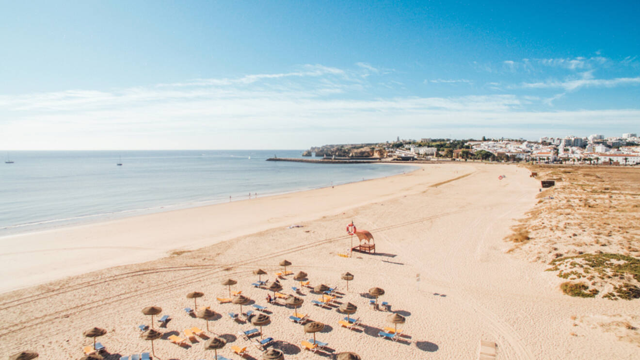 Spacious Meia Praia beach with scattered sun umbrellas, loungers, and a wide expanse of golden sand leading to a calm sea, with Lagos town in the distance