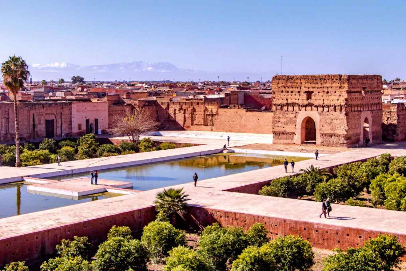View of El Badi Palace ruins in Marrakech, displaying historic red sandstone walls, reflective water pools, and a panoramic backdrop of the city skyline against the Atlas Mountains