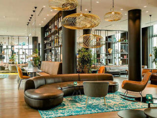 An upscale lounge at Motel One featuring a sleek, curved leather sofa, patterned rugs, and unique hanging lights in a modern space with a bookshelf-lined wall
