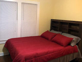 Simple bedroom setup with a red comforter, wooden headboard, and closed white blinds.