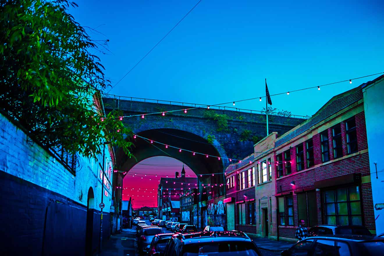 Twilight scene with a vivid blue sky over a street lined with parked cars, and string lights draped across an arching bridge