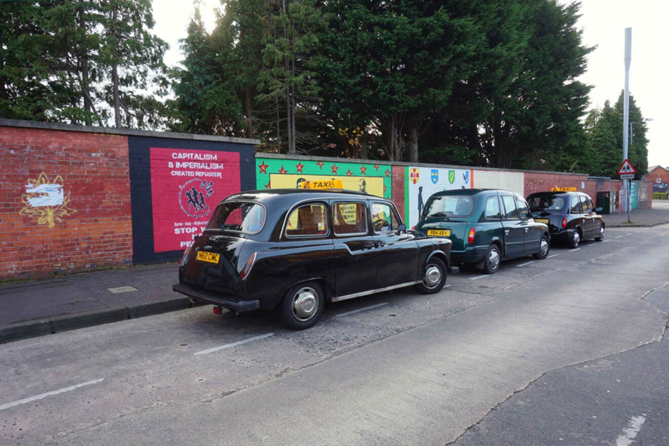 Row of traditional black cabs parked beside a political mural in Belfast, offering a glimpse into the city's unique taxi tours exploring historical and political sites.