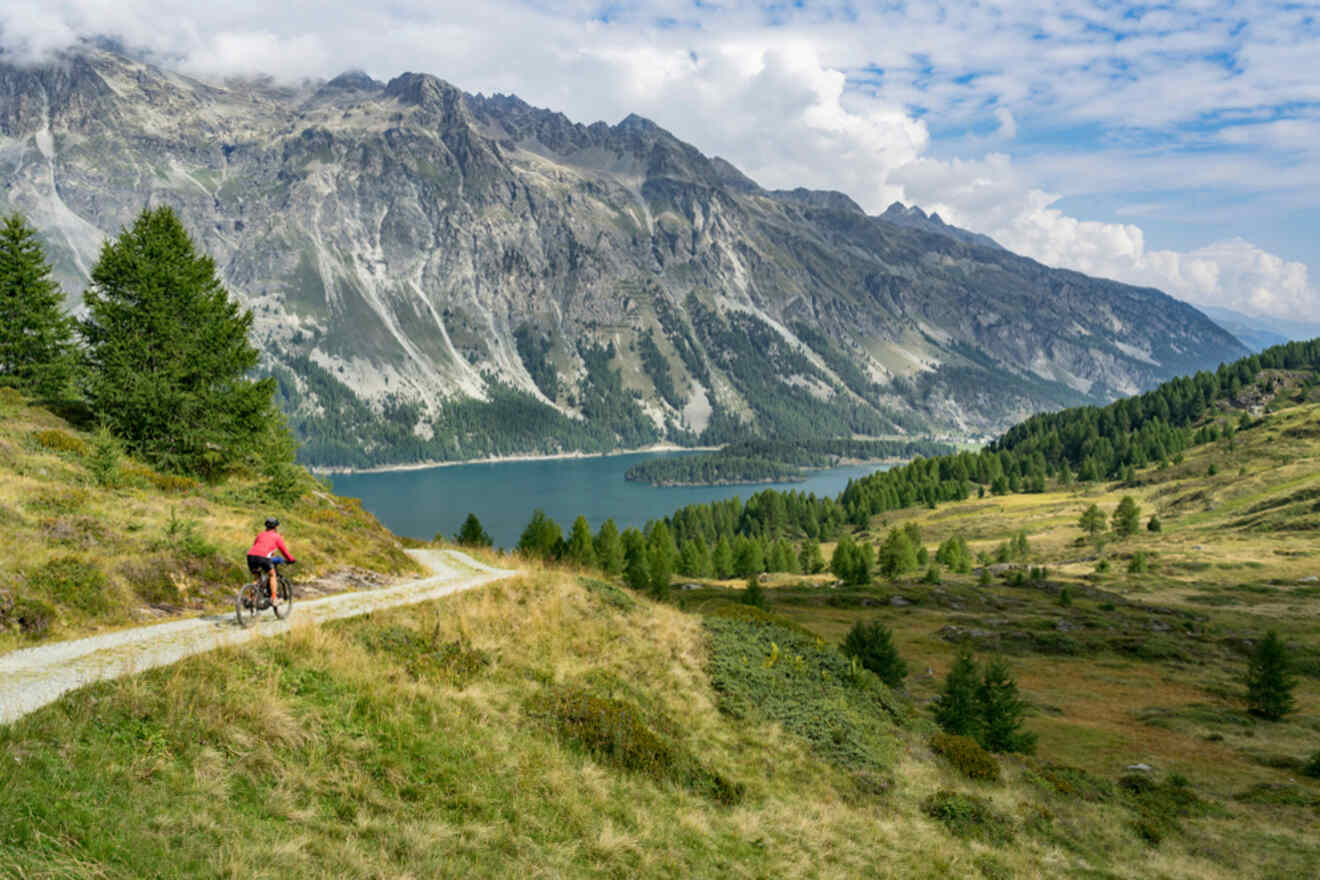 Cyclist in red attire riding along a mountain trail with panoramic views of a serene alpine lake and rugged peaks in the St. Moritz region