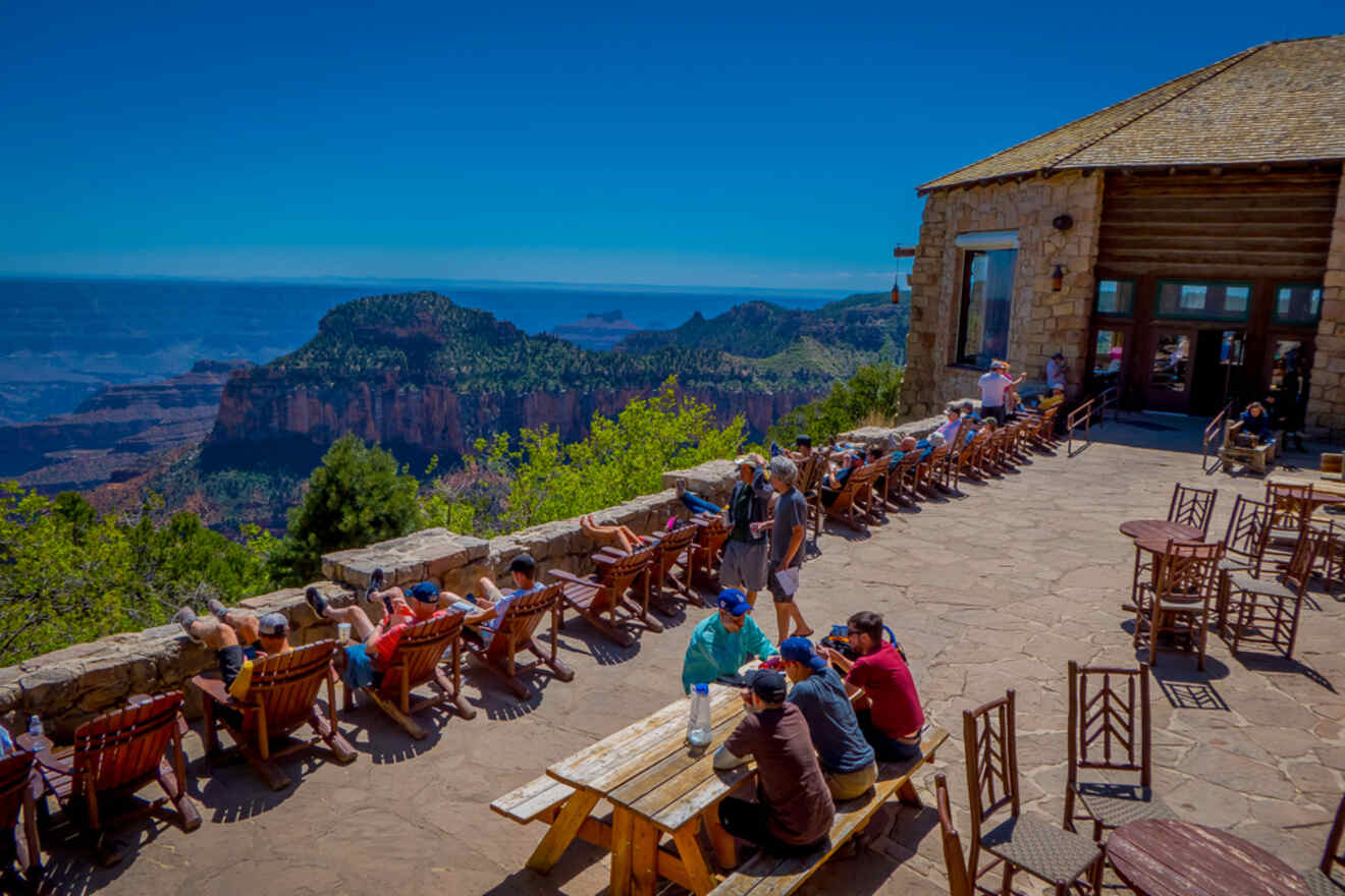 Outdoor seating area of a lodge with visitors enjoying the view over the Grand Canyon, a clear blue sky above and pine trees surrounding the terrace.