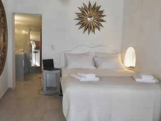 Minimalist bedroom with a white bed, sunburst mirror, and simplistic decor providing a tranquil Greek island vibe