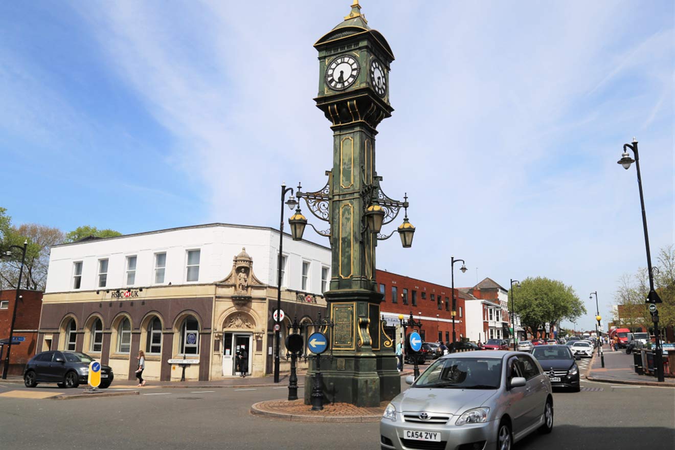 Ornate vintage clock tower standing at a busy intersection with cars and pedestrians, against a backdrop of a clear blue sky