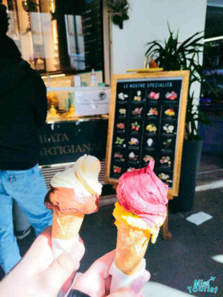 Close-up of two delicious-looking gelato cones, one with dark chocolate and vanilla, the other with raspberry, held in front of a gelateria's specialty sign
