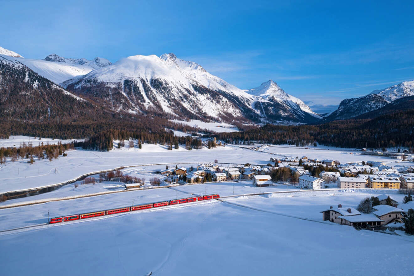 Red passenger train winding through the snow-blanketed valley of St. Moritz, with small clusters of buildings amidst vast fields and mountain ranges in the background