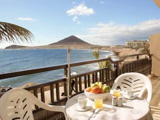 Table with two chairs set for breakfast on a balcony overlooking a beach, the ocean, and a mountain in the background under a partly cloudy sky.
