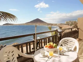 Table with two chairs set for breakfast on a balcony overlooking a beach, the ocean, and a mountain in the background under a partly cloudy sky.