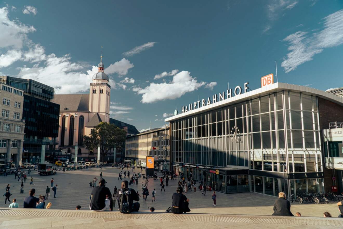The bustling front square of Cologne Central Station on a sunny day, with people sitting and walking around, and the station's mid-century modern architecture prominently visible