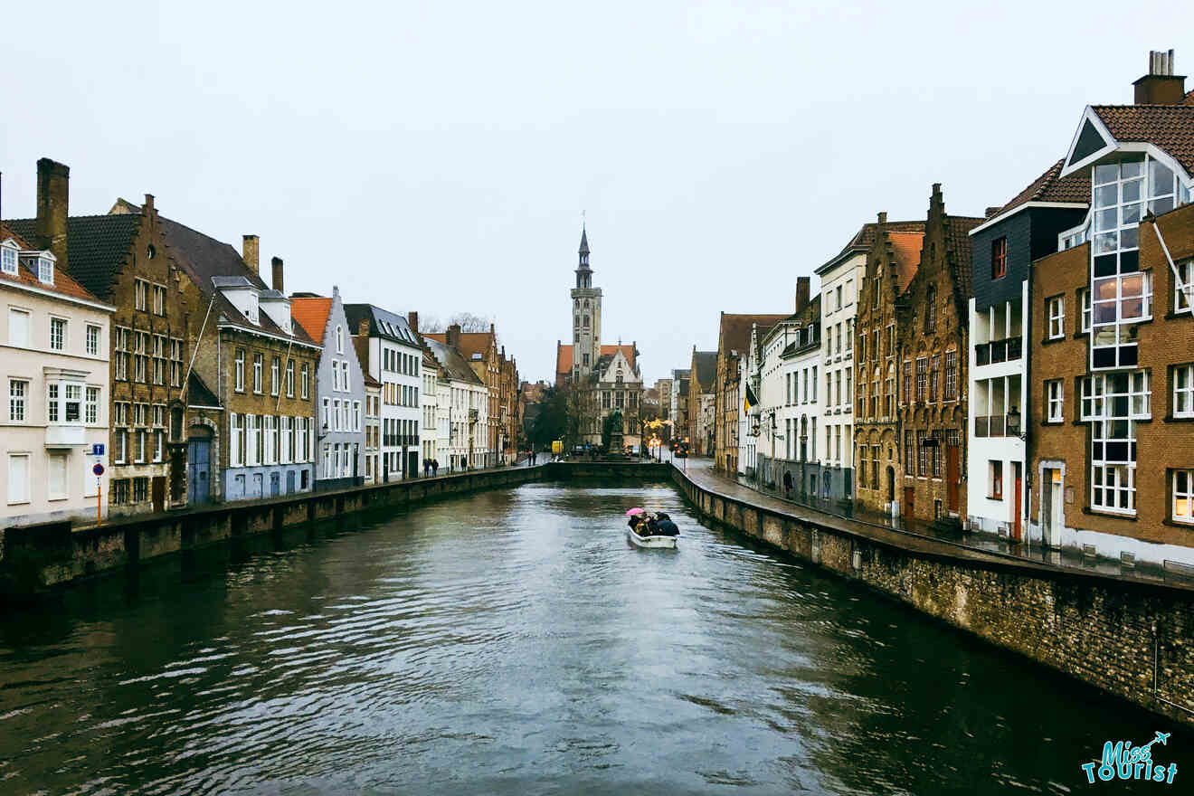"Cloudy day view of a canal in Bruges flanked by a row of traditional European houses, with a tower standing tall in the distance and a boat navigating the waterway