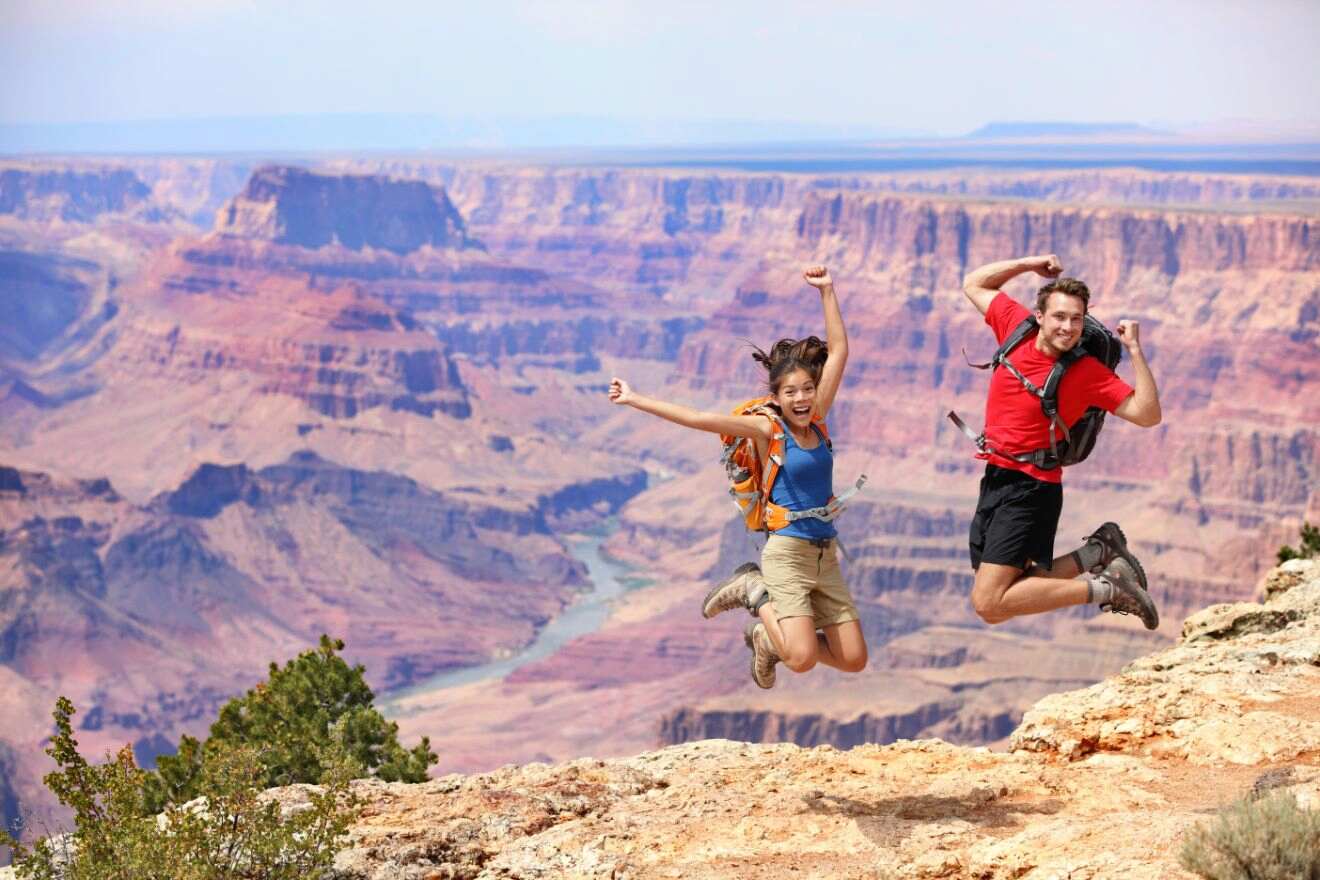 Two hikers joyfully leaping in the air on the edge of the Grand Canyon, showcasing the vast and colorful canyon landscape in the background.