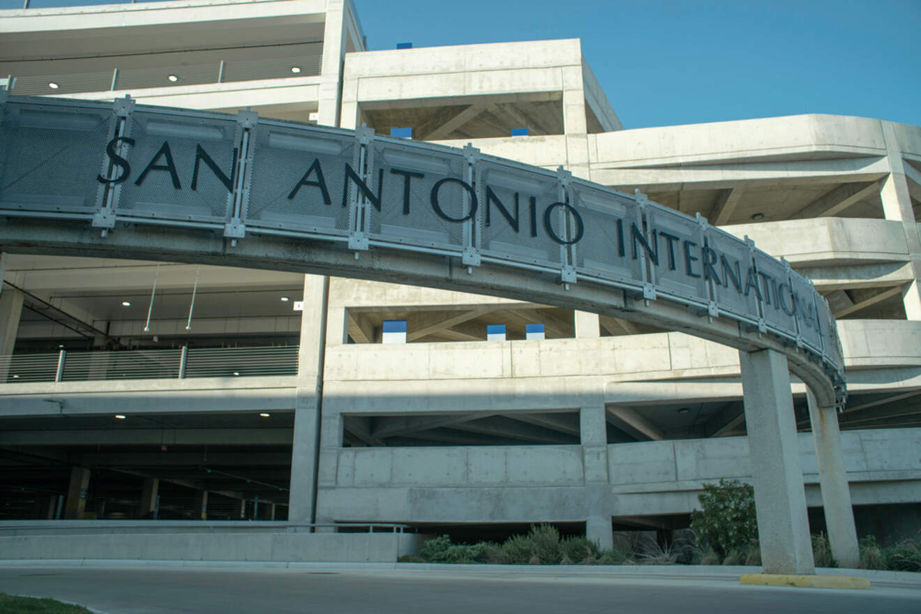 Entrance signage for San Antonio International Airport with a metallic finish on an overpass against a clear sky and the multi-level parking structure in the background.