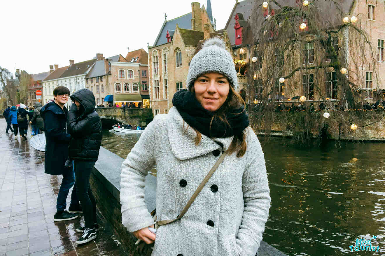 Author of the post in a cozy grey coat and knit hat smiling by the canal in Bruges, with historical buildings and festive lights in the background