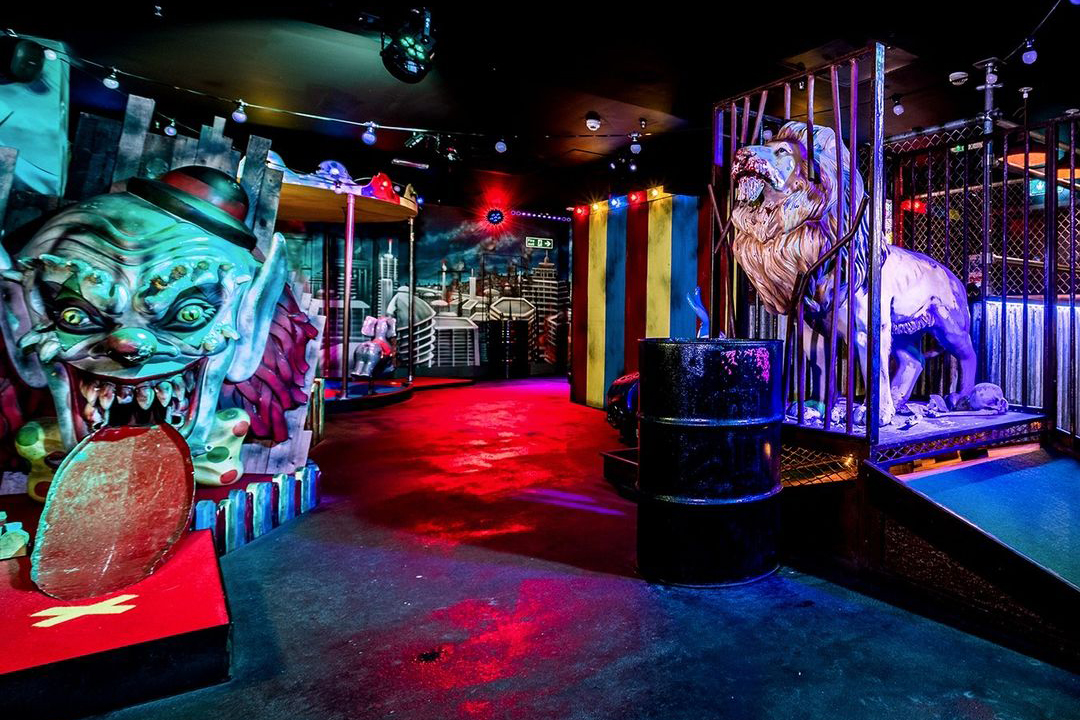 Indoor mini-golf course at Junkyard Golf Club with eccentric decor including a clown-faced obstacle, caged lion statue, and urban-themed artwork