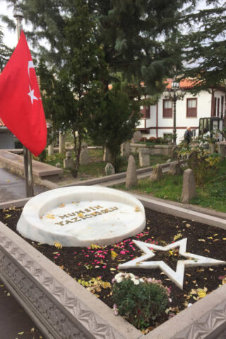 A tombstone and memorial decorated with Turkish flags, flowers, and an emblematic star and crescent in a serene, tree-lined graveyard.
