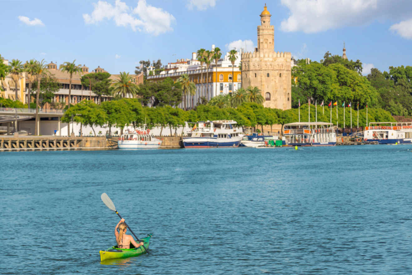 A person kayaking on the calm waters of River Guadalquivir with the historic Torre del Oro and lush trees in the backdrop, under a clear blue sky