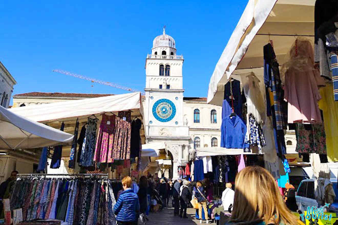 Vibrant market scene in Padua with stalls selling colorful textiles and clothes, with the iconic clock tower in the distance and shoppers milling about
