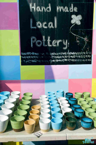 Assortment of handcrafted pottery displayed on a colorful stand with a handwritten sign