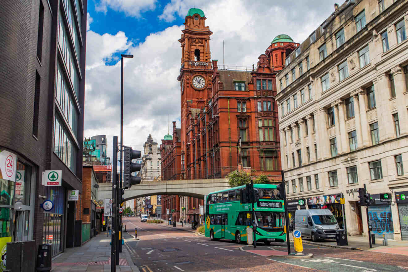 A view of a Manchester street with a red brick historic building with a clock tower, a green public bus in motion, and the pedestrian walkways on a cloudy day