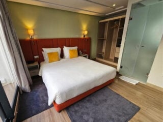 Guest room at DownTown Hotel Maârif Casablanca, with a rust-coloured upholstered bed, yellow cushions, open wardrobe and a glass-walled bathroom.